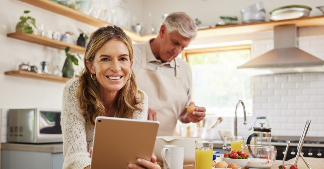 Woman 45+ years old looking to the camera with a table in her hand while her husband 50+ years old makes their breakfast